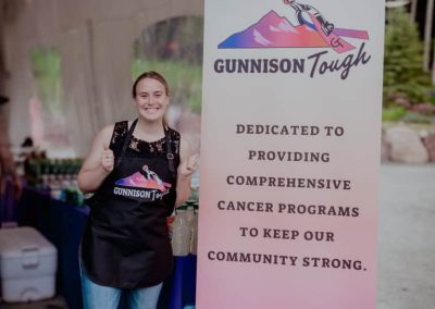A volunteer gives a thumbs up in front of a Gunnison Tough Sign that reads, "Dedicated to providing conprehensive cancer programs to keep out community strong."