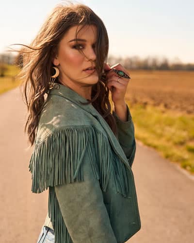 Mae Estes standing on a country road wearing a green leather jacket and beautiful jewelry.